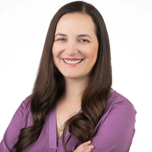 Woman with long dark hair in a purple top smiling with arms crossed.