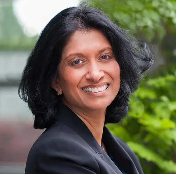 A woman with shoulder-length black hair, smiling, and wearing a black blazer, standing outdoors with greenery in the background.