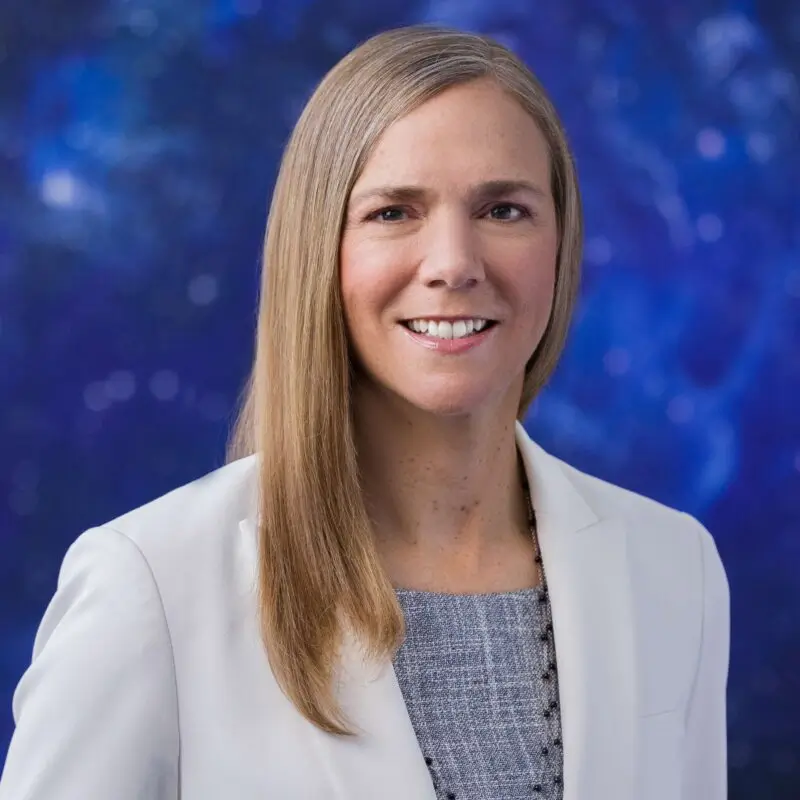 A woman with long straight blonde hair, smiling, wearing a light-colored blazer over a gray top, standing in front of a blue abstract background.