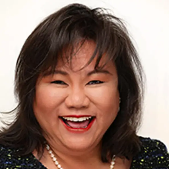 A woman with shoulder-length dark hair, smiling brightly, wearing a pearl necklace and a dark top, against a light background.