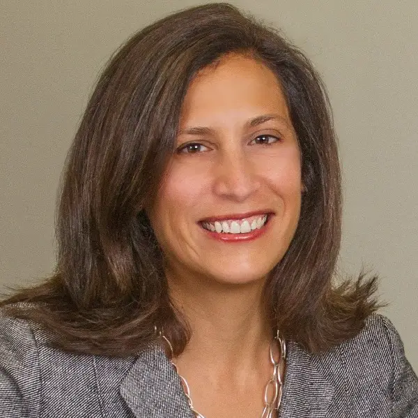 A woman with shoulder-length brown hair, smiling and wearing a gray blazer and a silver necklace, standing against a neutral background.