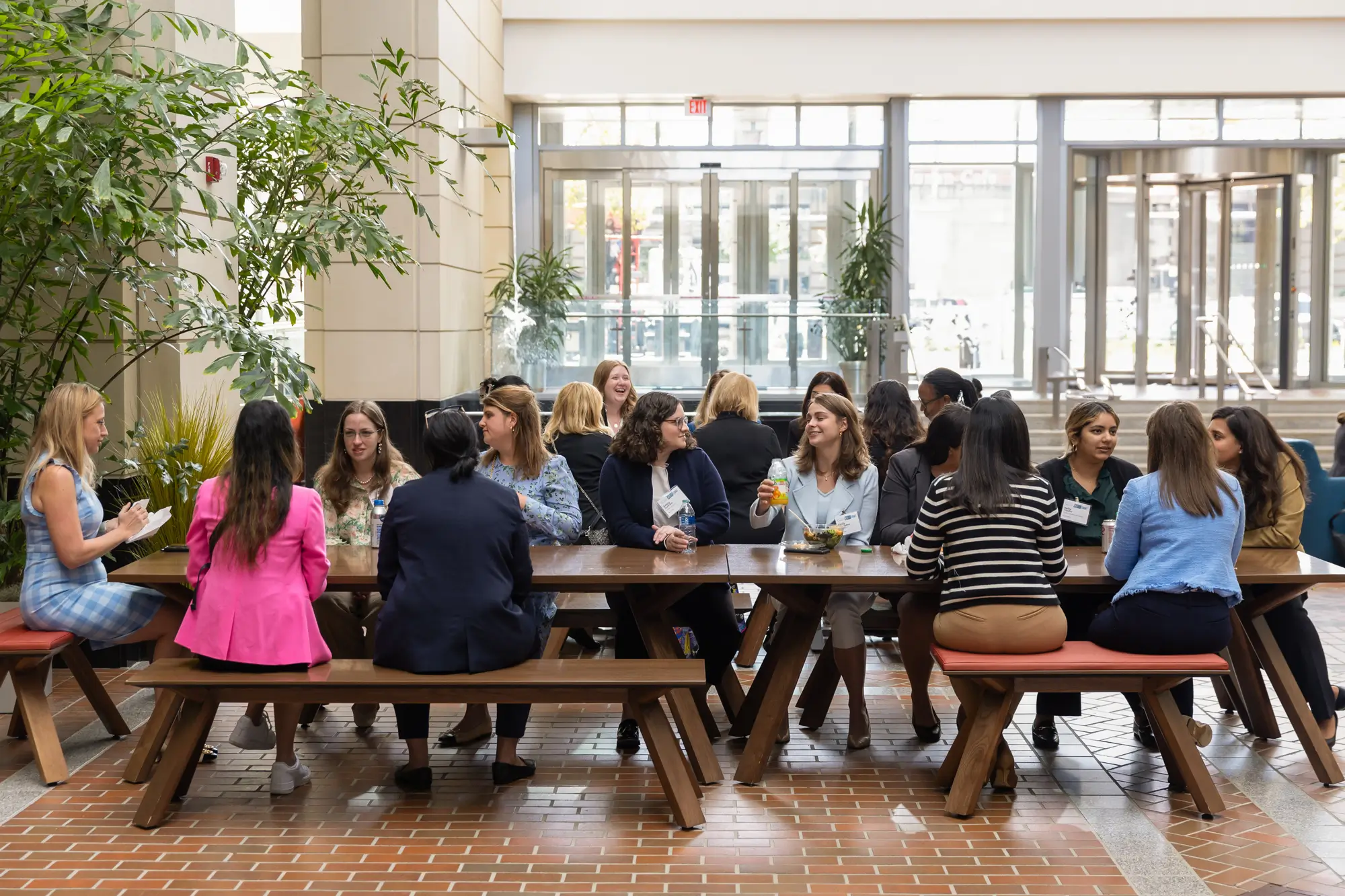 A group of people seated at a large wooden table in an indoor atrium, engaged in conversation and enjoying drinks and food. The atrium has large windows, plants, and natural light.