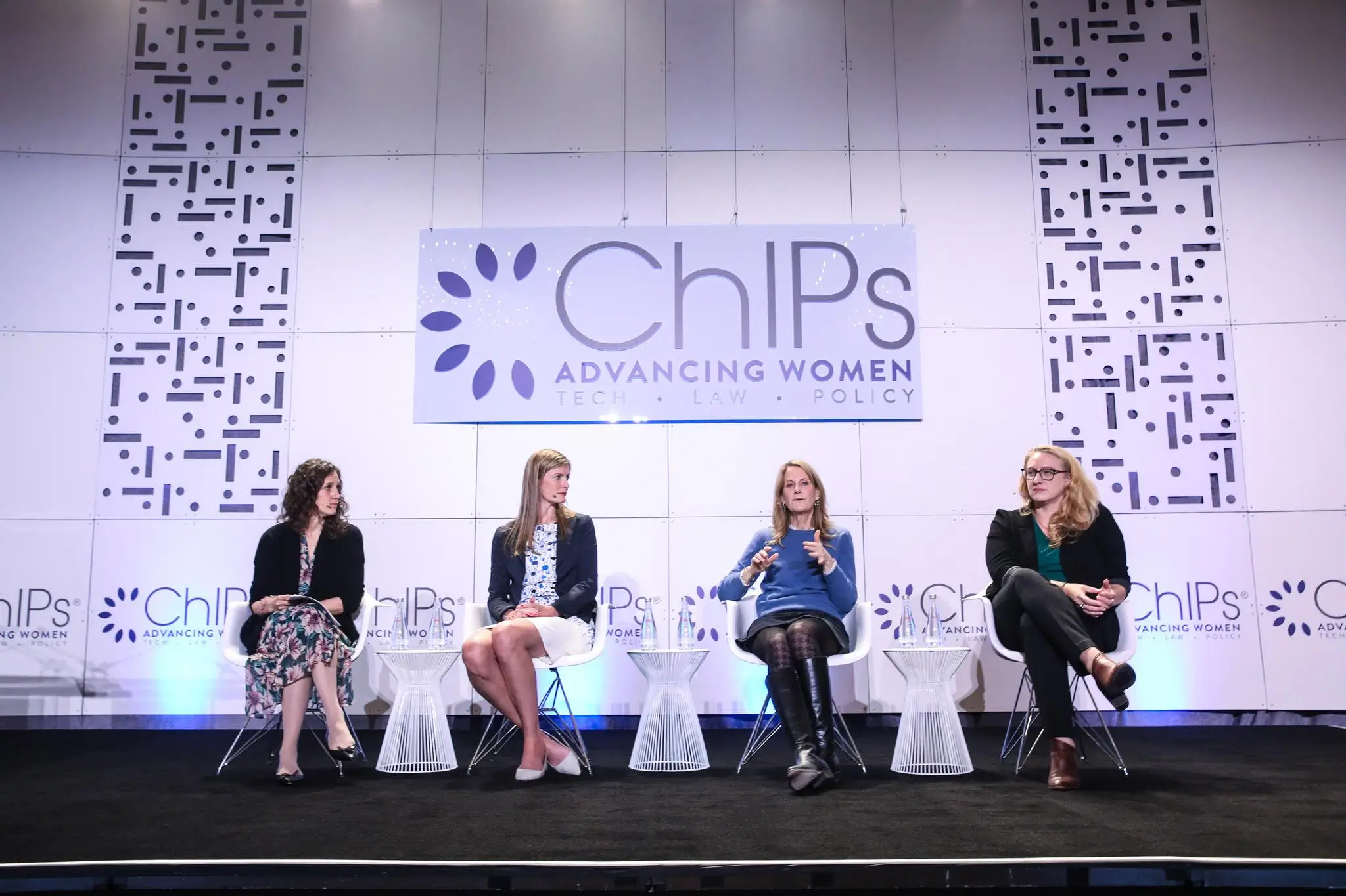 Four women seated on stage at the ChIPs Global Summit, engaged in a panel discussion. The backdrop displays the ChIPs logo and the text 'Advancing Women in Tech, Law, Policy.' Each woman is seated in a chair with microphones and water glasses on small tables in front of them.