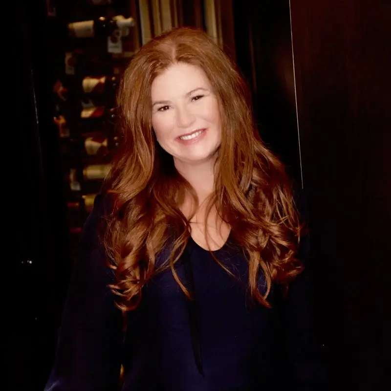 A woman with long, wavy red hair, smiling and wearing a dark top, standing indoors with a wine rack in the background.