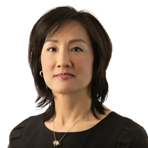 A woman with short dark hair, wearing a black top, gold hoop earrings, and a pendant necklace, standing against a white background.