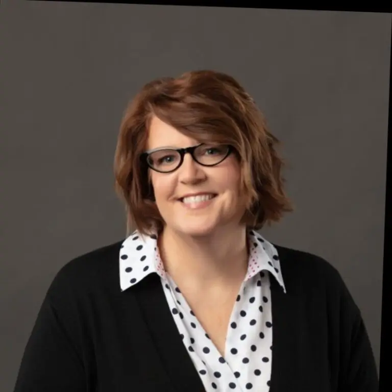 Woman with short auburn hair and glasses, dressed in a black sweater over a white polka dot blouse.