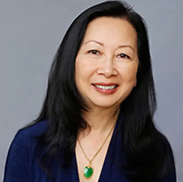 A woman with long dark hair, smiling, wearing a dark blue top and a necklace with a green pendant, against a gray background.