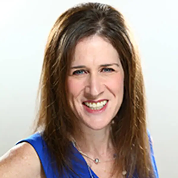 A woman with long straight brown hair, smiling and wearing a bright blue sleeveless top, standing against a light background.