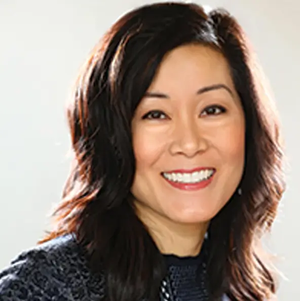 A woman with shoulder-length dark hair, smiling, and wearing a dark textured top against a light background.