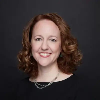 A woman with shoulder-length curly red hair, smiling and wearing a black top and a layered necklace, standing against a dark background.