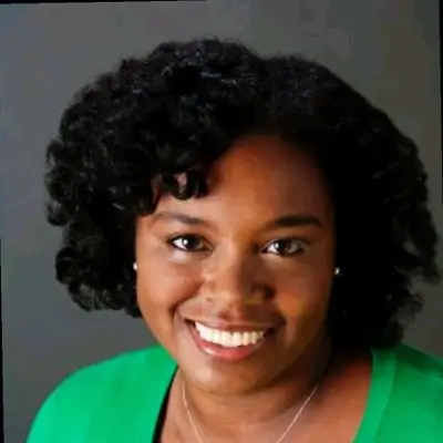 A woman with curly dark hair, smiling, and wearing a bright green top and a necklace, standing against a dark background.