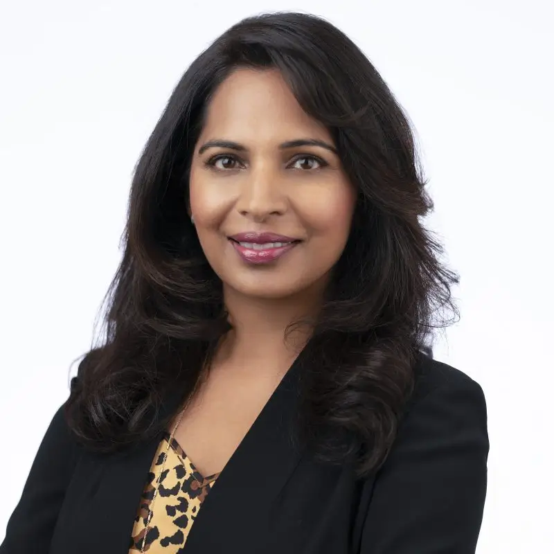 A woman with long dark hair, smiling, and wearing a black blazer over a leopard print top, standing against a white background.