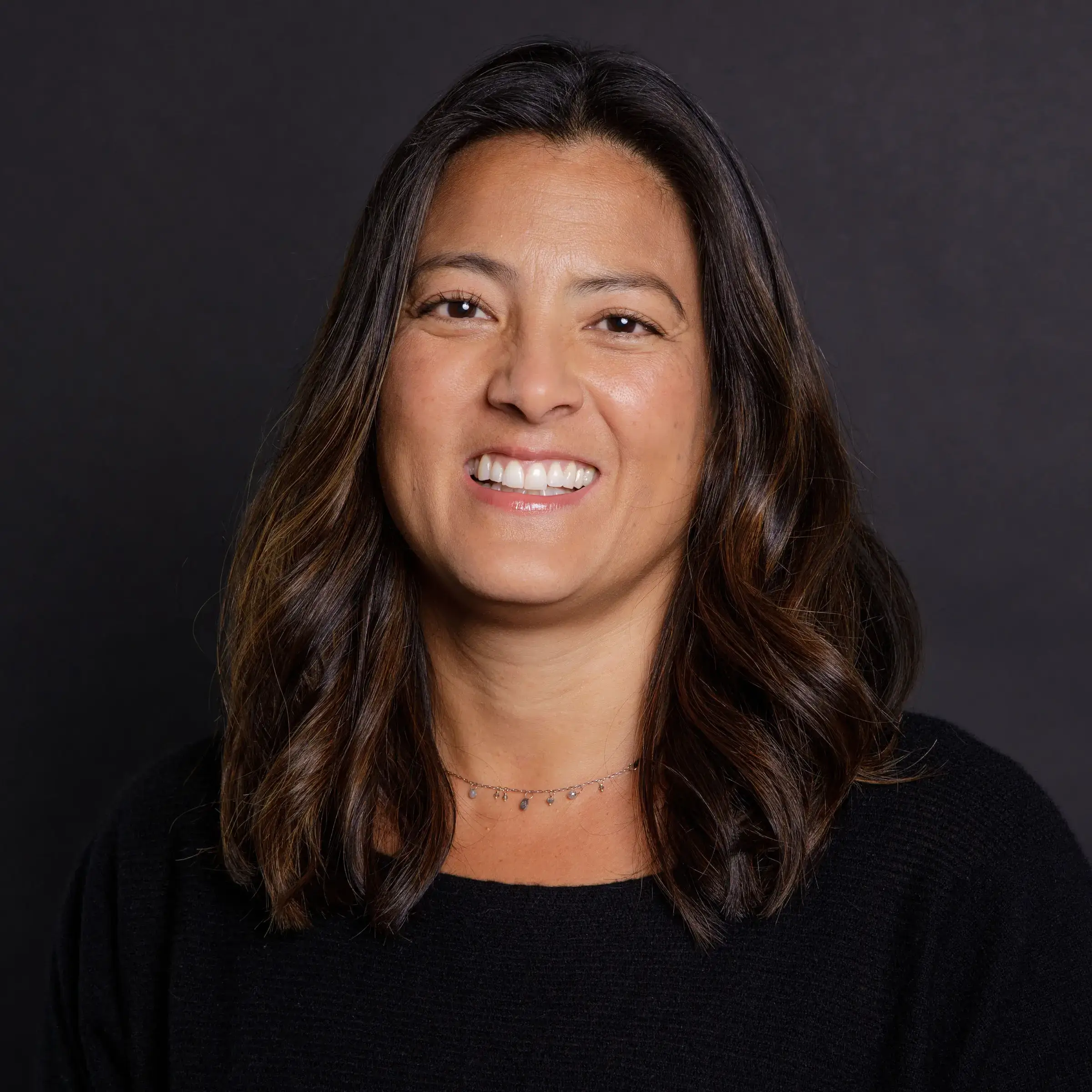 A woman with shoulder-length dark hair, smiling, and wearing a black top and a delicate necklace, standing against a dark background.