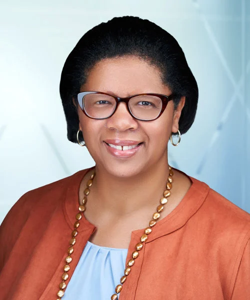 A woman with short dark hair, wearing glasses, an orange blazer, and a beaded necklace, smiling confidently in front of a light blue background.