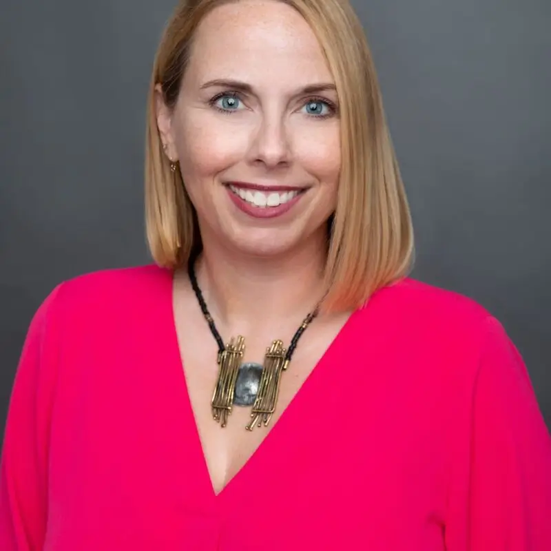 Portrait of a smiling woman with shoulder-length blonde hair, wearing a bright pink top and a statement necklace, against a gray background.