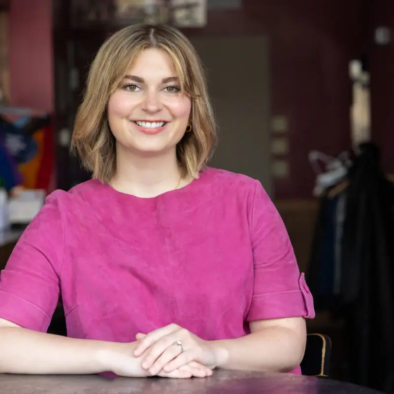 Portrait of a smiling woman with shoulder-length blonde hair, wearing a bright pink top, seated at a table in a warmly lit indoor setting.