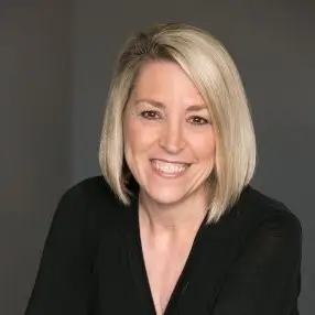 Headshot of a smiling woman with short blonde hair, wearing a black top, against a neutral gray background.