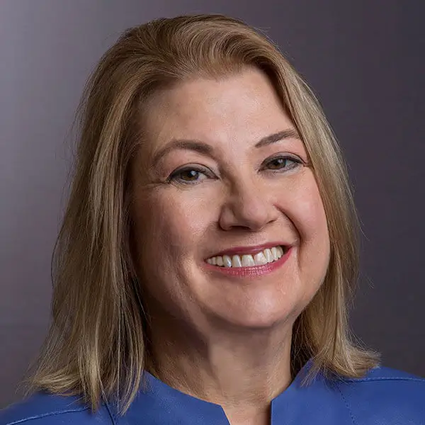 Portrait of a smiling woman with shoulder-length blonde hair, wearing a blue top, against a dark background.