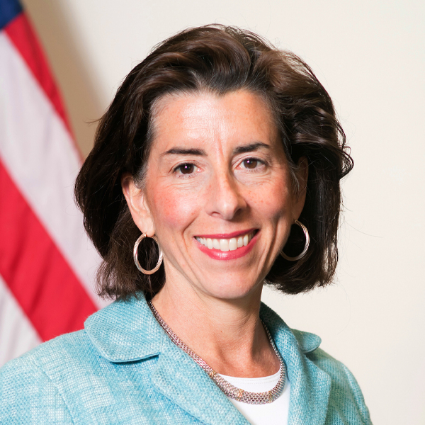 Gina Raimondo, smiling and wearing a light teal blazer with a silver necklace, stands in front of an American flag background.