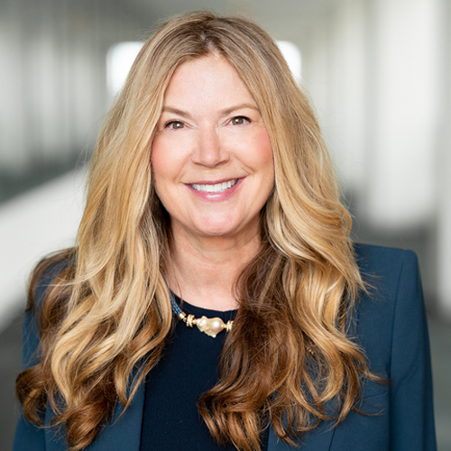 Kathi Vidal, smiling and wearing a dark teal blazer with long wavy blonde hair, standing in a professional indoor setting.