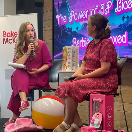 Two women seated on stage, one holding a microphone and speaking, both dressed in pink. Behind them, a large screen displays the title 'The Power of IP Licensing: Barbie Unboxed.' Various Barbie-themed items and a beach ball are placed near the stage, with a Baker McKenzie logo in the background.