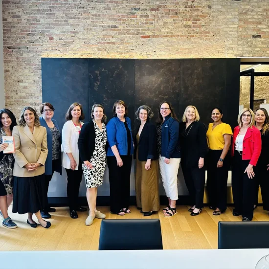 A group of twelve women standing in a line in a professional setting, smiling at the camera. They are dressed in a variety of business and casual attire, standing in front of a brick wall and dark panels.