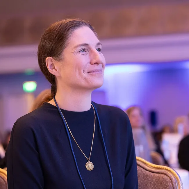 A woman with a ponytail, wearing a dark top and a gold pendant necklace, smiles and looks attentively ahead while seated at the Global Summit, with a softly lit background.