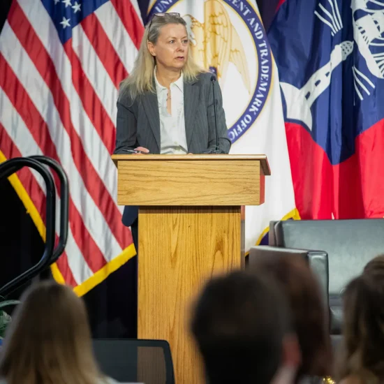 Woman in gray suit at a podium in front of a US flag.